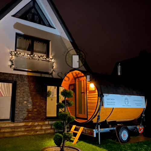 Eine weihnachtlich beleuchtete Fasssauna bei Nacht im Garten vor einem beleuchteten Haus