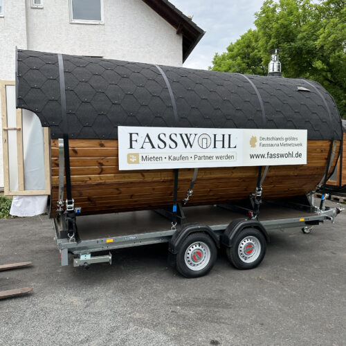 Eine Fasssauna mit großem "Fasswohl" Schild auf einem Anhänger